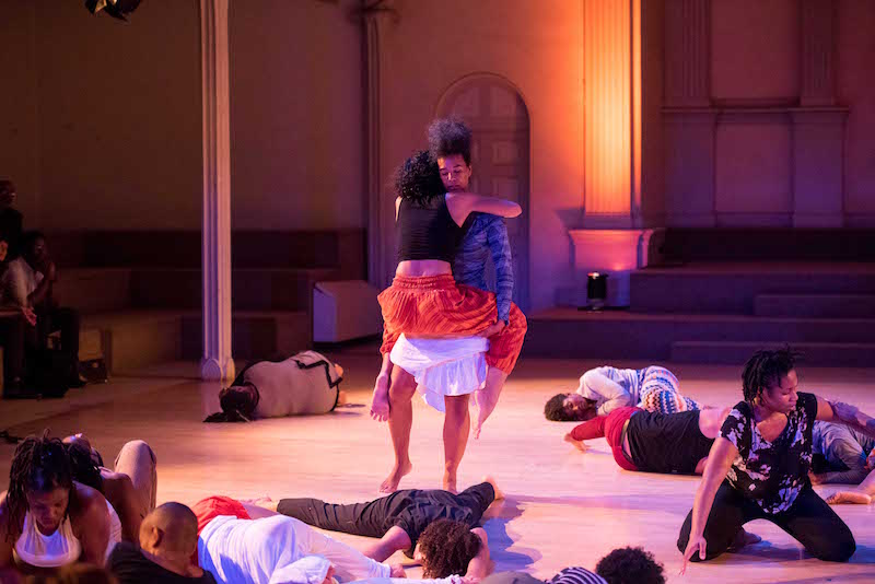 A woman holds another woman in red pants like a baby. Other dancers lay on the ground.